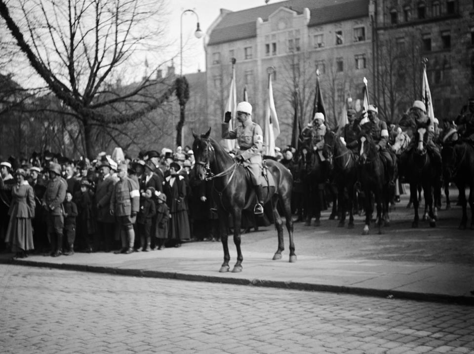 Marsalkka Mannerheim ratsunsa selässä. Mannerheimin takana joukko sotilaita hevosten selässä Suomen liput vierellä. Ihmisiä seuraamassa.