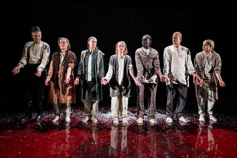 kuusi ihmistä seisoo rinnakkain käsivarret eteen ojennettuina. Kaikilla on käsivarressa tatuointi ja he katselevat tatuointeja ja toisiaan ihmetellen