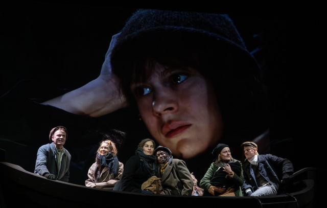 kuusi ihmistä istuu veneessä. Yhden kasvot on projisoitu taakse suurina