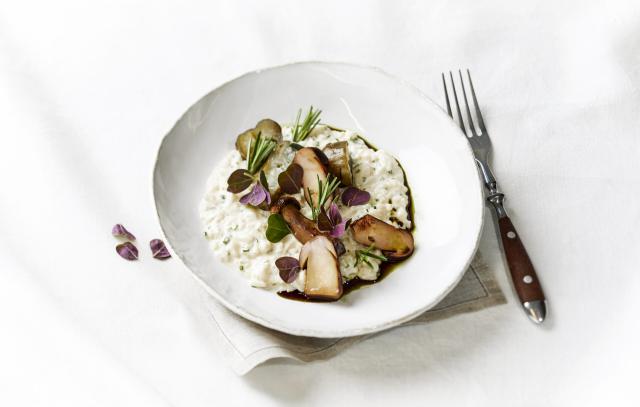 risottoannos valkoisella lautasella valkoista pöytäliinaa vasten, risoton päällä on paahdettua viipaloituja sieniä sekä yrttejä