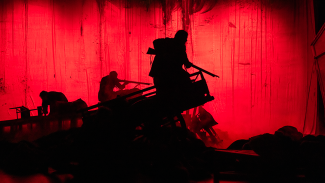 punainen näyttämökuva, jossa hahmotettavissa vain kolme tummaa hahmoa. Etualalla olevista kaksi pitää kivääriä käsissään ja katsoo samaan suuntaan. Kolmas on kyyristynyt.