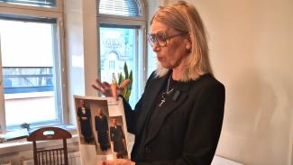 mustassa asussa oleva vaaleahiuksinen nainen esittelee paperiarkkia, jossa on kolmen eri henkilön kuvat.