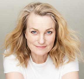 Keski-ikäinen nainen katsoo suoraan kohti kasvoillaan pehmeä hymy. Hänellä on vaaleat olalle ylettyvät hiukset ja valkoinen t-paita.